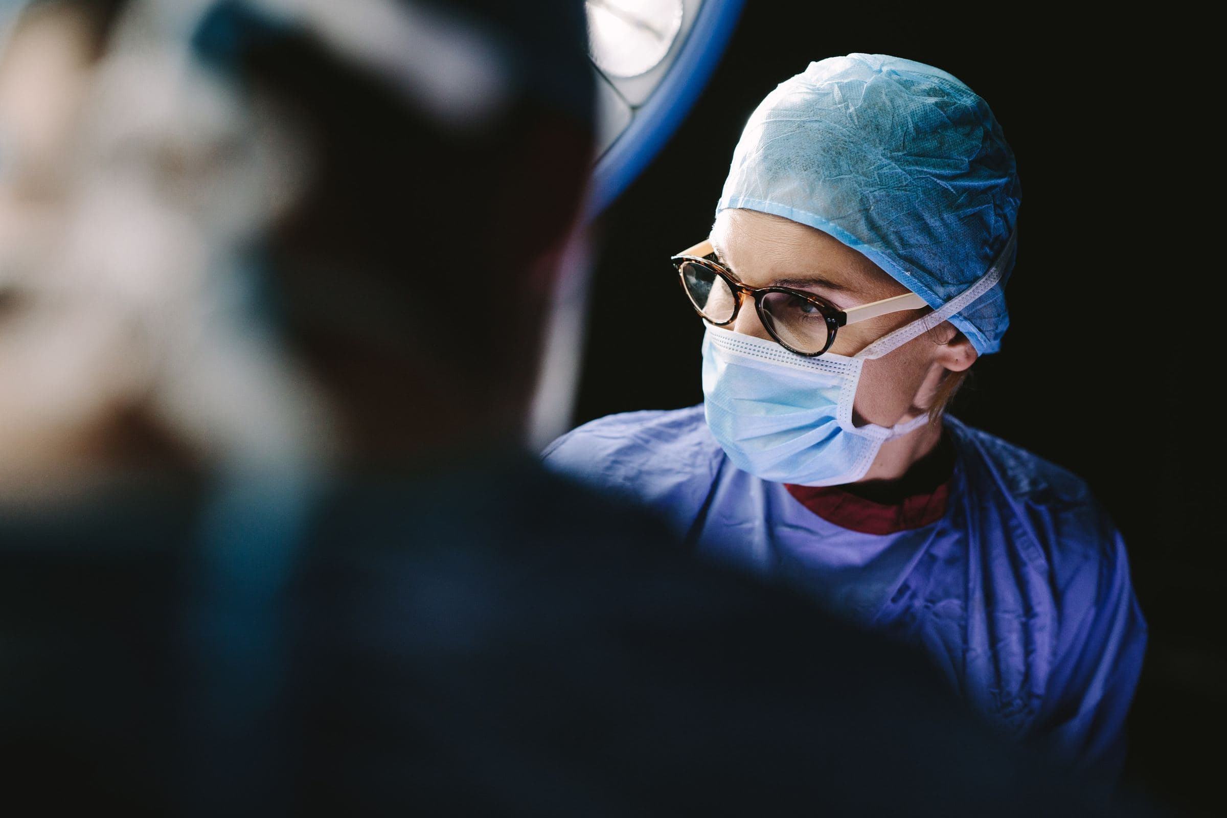 A clinician in a surgical cap, mask and glasses, seen past a colleague’s shoulder